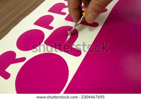 The hand of a man with a stationery knife, selects a vinyl film after cutting on a plotter.Sampling of the film in the production of avtovinil. Cutting a pink sticker.