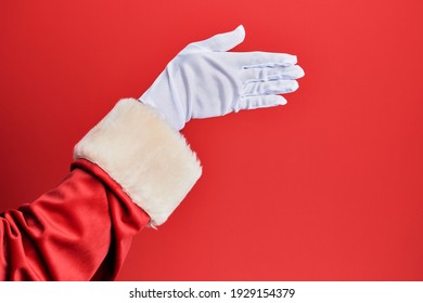 Hand Of A Man Wearing Santa Claus Costume And Gloves Over Red Background Stretching And Reaching With Open Hand For Handshake, Showing Back Of The Hand 