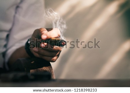 Hand of a man in white basic shirt sitting in the shadow and smoking cigar.Man spent his time with a good cigar.Blurred image of small gun at foreground.Concept of mafia or god's father or big guy.