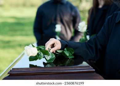 Hand of mature woman in mourning attire putting white rose on top of closed coffin lid while standing in front of camera against other people