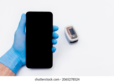 The Hand Of A Medical Doctor In A Latex, Rubber Glove Holds A Mobile Phone Or Smartphone With A Screen Against The Background Of The Pulse Oximeter.