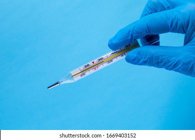 Hand in a medical glove holds a thermometer, a thermometer close-up on a blue background. A thermometer in the hand of a medical professional shows high body temperature which confirms an outbreak