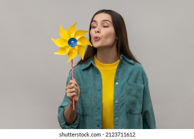 Hand mill. Portrait of happy beautiful young adult woman blowing at paper windmill, pinwheel toy on stick, wearing casual style jacket. Indoor studio shot isolated on gray background.
