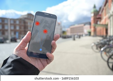 Hand With Mobile Phone And Coronavirus Tracking App Against The Spread Of The Pandemic, But Controversial When It Comes To Protection Of Personal Data, Blurry City Background With Copy Space