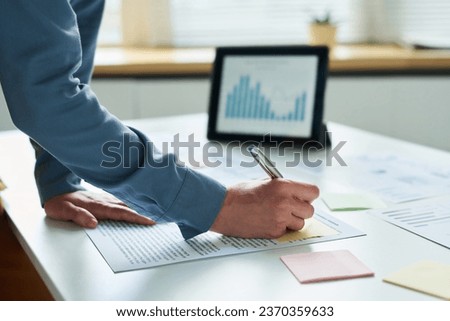 Hand of modern businesswoman in blue shirt making notes on notepaper after reading contract or some other financial document in office