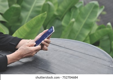 Hand of old woman hold her smartphone sitting and relaxing outdoor in the garden. 