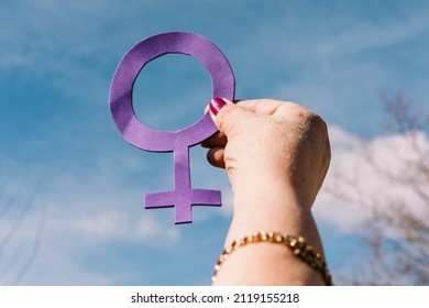 Hand Of An Older Woman With Purple Nails, With The Sky In The Background, Holding The Female Symbol In Purple. Concept Of Women's Day, Empowerment, Equality, Inequality, Activism And Protest.