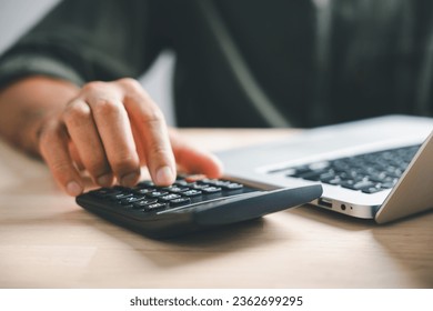 Hand on calculators, aiming for home refinance. Wooden house model, buy or rent note on desk. Concept of saving to buy property, wise mortgage payment. Tax, credit analysis for financial benefit.