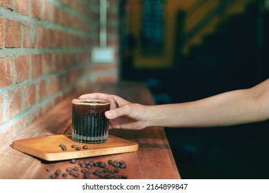 Hand on a Glass of ice coffee, americano in the morning over a red bricks wall background, and coffee beans on a wood counter bar table. Cold brew refreshment summer drink with copy space. Cafe idea.