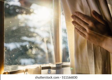 Hand Opens Curtain On The Wooden Cottage House Window. Bright Sunlight Shines Through. Beautiful Snowy Winter Morning Light Behind The Chalet Window.