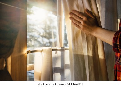 Hand Opens Curtain On The Wooden Cottage House Window. Bright Sunlight Shines Through. Beautiful Snowy Winter Morning Light Behind The Chalet Window.