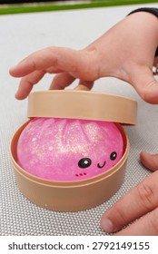 A hand opens a small beige container to reveal a pink glittery dumpling squishy toy with a smiling face, perfect for stress relief and play