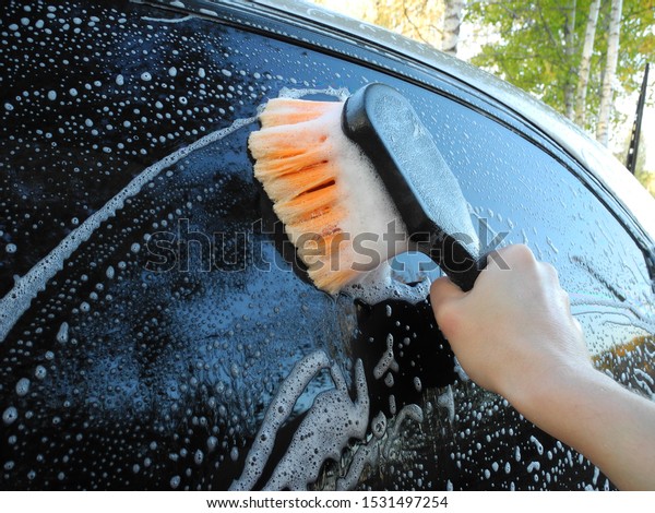 Hand with orange brush washes car glass with foamy\
detergent on background of birches. Home car wash.                 \
            