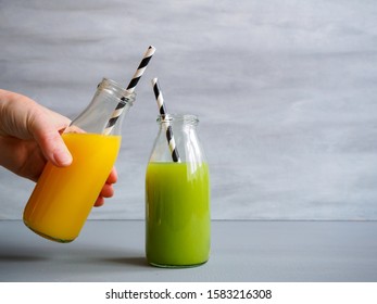 Hand With Orange Juice In A Glass Bottle And Green Vegetable Juice On A Gray Background, Drink To Go