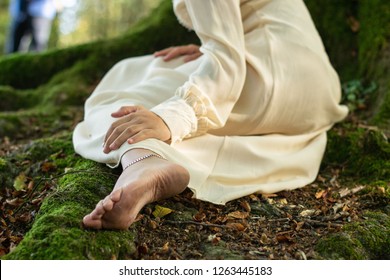 Hand over foot of a barefoot woman on a white dress, sitting on a tree root
