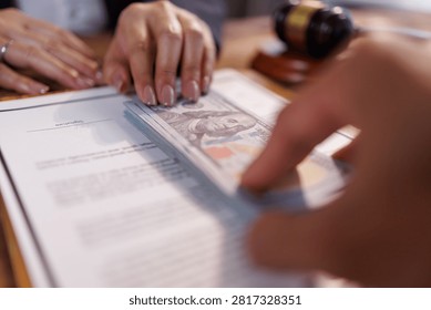 Hand passing a stack of hundred dollar banknotes over a contract document, symbolizing illegal payments, bribery, judicial corruption, and the subversion of legal agreements