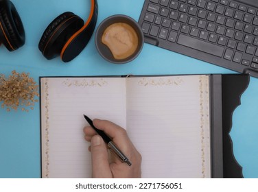 hand of person taking notes in notebook or journal adorned flowers on edge with a coffee keyboard dried flower and headphones with office light on blue or turquoise desk with text pear space