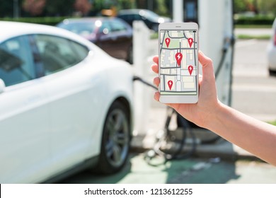 A hand with a phone on the background of a charging station for electric cars. On the screen is a map with the location of the charging stations.