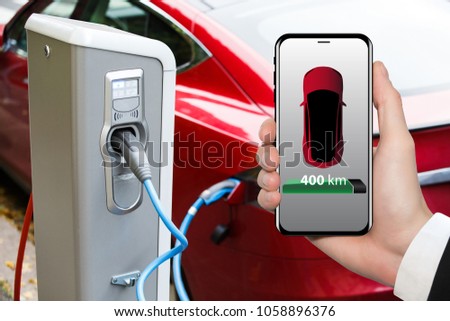 Hand with phone on a background of electric car charging point. On a device screen indicator of power reserve. 
