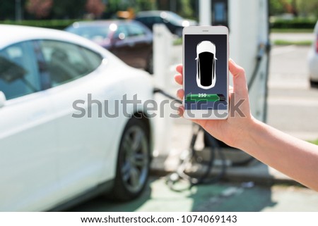 Hand with phone on a background of electric car charging point. On a device screen indicator of power reserve. 