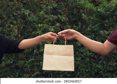 Woman’s Hand Pick Up Taking Empty Craft Brown Recycled Paper Shopping Bag From Man With Outdoor Green Leaves Nature Hedge Background. Delivery Paper Bag Mockup For Branding Design Concept