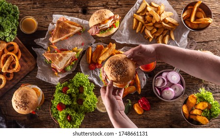 Hand Picking Up A Burger From A Top View Of A Table Full Of Fast Food, Including Hamburgers, Potato Chips, Club Sandwich, Chicken Nuggets And Onion Rings On A Rustic Table.