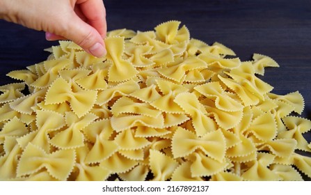 Hand Picking a Piece of Farfalle or Bow-tie Pasta from Its Pile on Black Wooden Background