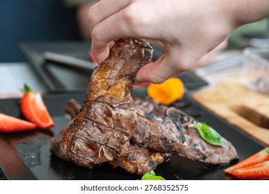 a hand picking up a stack of juicy grilled lamb chops with distinct grill marks from a square black plate beautifully garnished with fresh red strawberries and green basil leaves