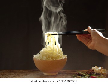 Hand Picking Up Tasty Steaming Noodles With Chopsticks In Wooden Bowl On Wooden Background. Selective Focus Asian Dish On Table. Junk Food Concept.. 