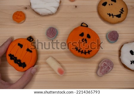 A hand picks up an orange pumpkin-shaped cookie from a light wood board littered with Halloween cookies, candies and sweets. At a spooky children's party. White, yellow and brown colors. Wallpaper