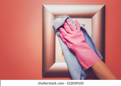 Hand In Pink Protective Glove Wiping Picture Or Photo Frame On The Wall From Dust With Rag. Early Spring Cleaning Or Regular Clean Up. Maid Cleans House.