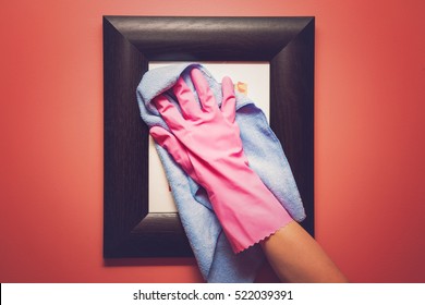 Hand In Pink Protective Glove Wiping Picture Or Photo Frame On The Wall From Dust With Rag. Early Spring Cleaning Or Regular Clean Up. Maid Cleans House.