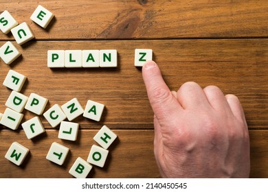 A hand places a piece with the letter Z next to the word PLAN; around it other letters appear in disarray. Concept of planning for the future, resilience and constancy. Background of wooden boards.