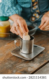 Hand placing solid ingredient into metal cup on digital scale during homemade preparation on rustic wooden table