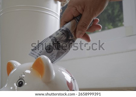 Hand placing stack of dollar bills into white piggy bank, symbolizing saving money and financial planning. scene is set indoors with natural light coming through window, creating calm atmosphere