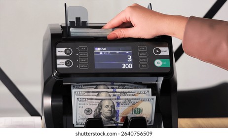 Hand placing US dollars into an electronic money counter. A close-up view of a person inserting a stack of US dollar bills into a digital currency counting machine. - Powered by Shutterstock - Get 15% off with code: PIKWIZARD15