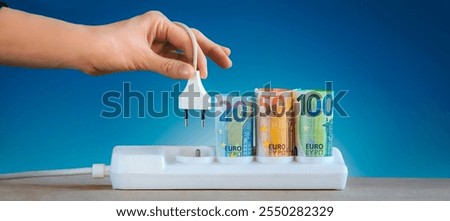 hand plugging an electric cable into a power . Euro money paper banknotes plugged into a white power strip Increasing cost of electricity for business users and residential customers. Rise in electric