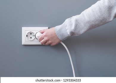 Hand plugging in an electric cord into a white plastic socket or  european wall outlet on grey plaster wall. Closeup of a woman's hand inserting an electrical plug into a wall socket. Daylight. 