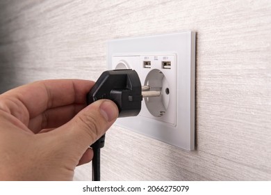 Hand Plugging In An Electric Cord Into A White Plasticmodern Socket Or European Wall Outlet On Grey Wall. Closeup Of A Man Hand Inserting An Electrical Plug Into A Wall Socket.