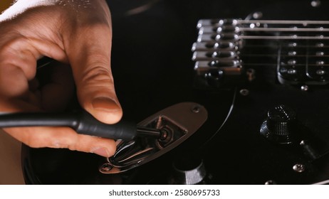 A hand plugging an instrument cable into an electric guitar's input jack, with bridge saddles and volume knob visible on the glossy black body. The lighting creates subtle reflections