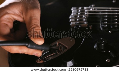 A hand plugging an instrument cable into an electric guitar's input jack, with bridge saddles and volume knob visible on the glossy black body. The lighting creates subtle reflections