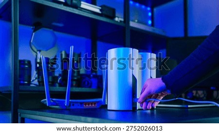 A hand plugs an Ethernet cable into a sleek mesh Wi-Fi unit, with a traditional router and camera equipment visible in the background
