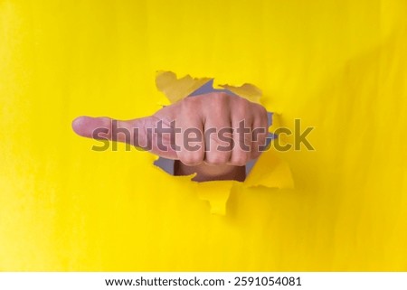 A hand pointing through a torn yellow paper, symbolizing direction, command, or accusation.