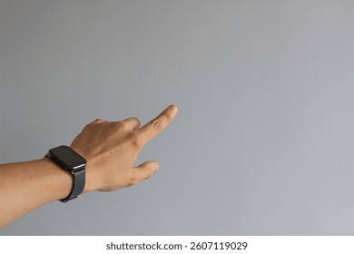A hand points forward with an extended index finger, wearing a sleek black smart watch. Set against a plain light gray wall, ideal for modern tech and directional themes