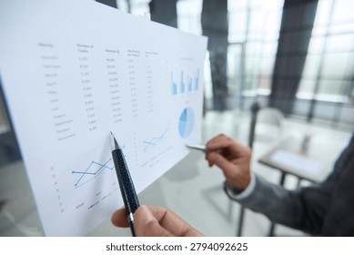 Hand points to graph, showing data trends. Person analyzes figures with focus, using pen. Business meeting, planning strategy, or review. Professional setting, modern office with glass
