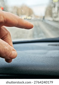 A hand points at the road while driving inside a car on a dirty street during daylight. Trees are visible in the background with traffic lights ahead.