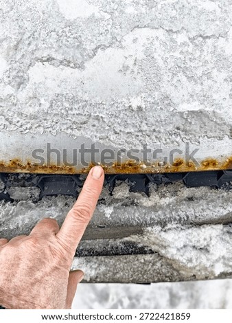 Hand points at rust and corrosion on car hood edge. Winter dirt and road salt are visible, showing vehicle maintenance needs.