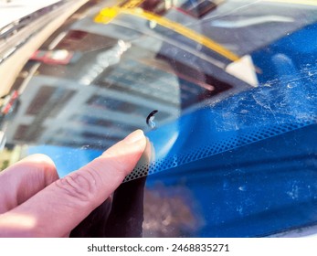 A hand points to a small chip on the driver-side window of a parked car, highlighted in bright daylight