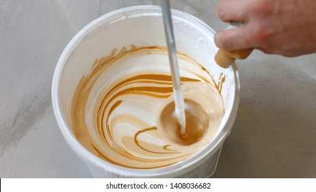 Hand Pouring Brown Liquid Into White Paint. Person Mixing White Paint In Bucket. Home Renovation Concept.