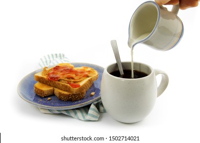 A Hand Pouring Cream Into A Cup Of Coffee With A Peanut Butter And Jam On Toast Isolated On White